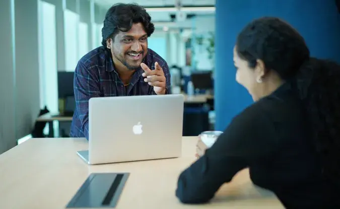 A male employee sits at a laptop, laughing with a female coworker in a conference room.