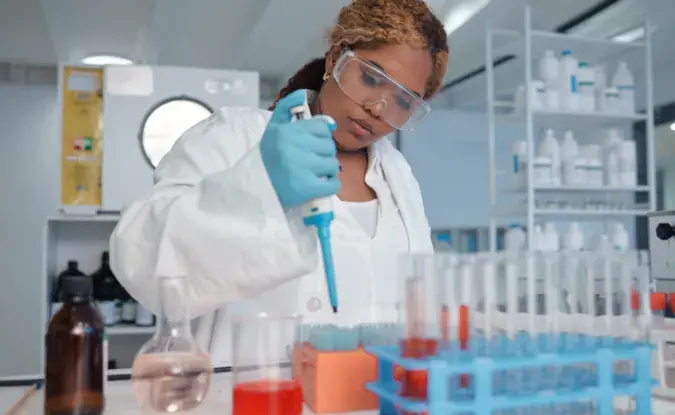 Lab worker in white coat and safety glasses uses a special tool to fill a tray of glass vials.