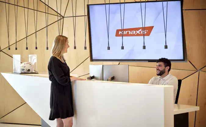 A woman in a black dress smiles as she stands at the Kinaxis reception desk in Ottawa, and a man sits at the reception desk to welcome her. The red Kinaxis logo is displayed on the wood wall behind the desk.